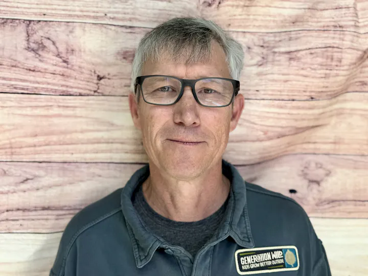 A man with glasses stands in front of a wooden backdrop, wearing a jacket with a patch that says "Generation Wild."
