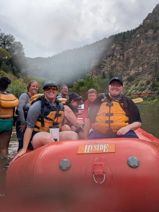 A group of people in orange life vests sit in a red raft by a river, surrounded by trees and mountains on a cloudy day.