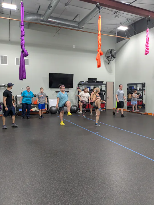 A group of people engaged in a fitness or dance class, with colorful fabrics hanging from the ceiling and exercise equipment around.