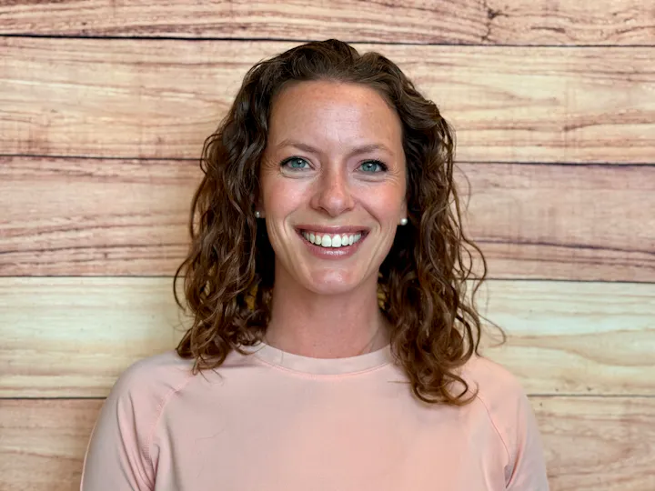 The image shows a smiling woman with curly hair, wearing a pale pink top, against a wooden backdrop.