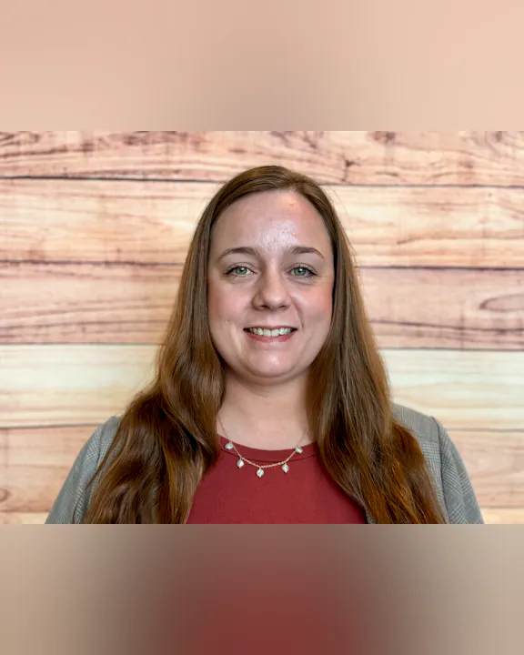 A woman smiles in front of a wooden backdrop, wearing a red top and a necklace, with long hair and a friendly demeanor.