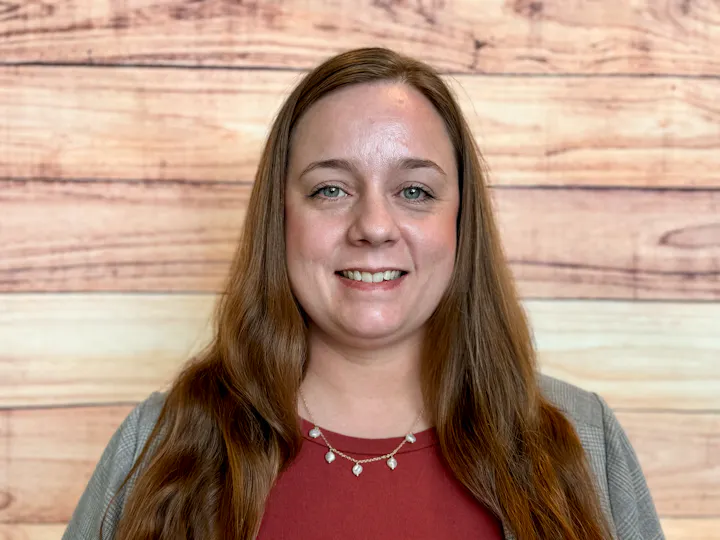 A woman smiles in front of a wooden backdrop, wearing a red top and a necklace, with long hair and a friendly demeanor.
