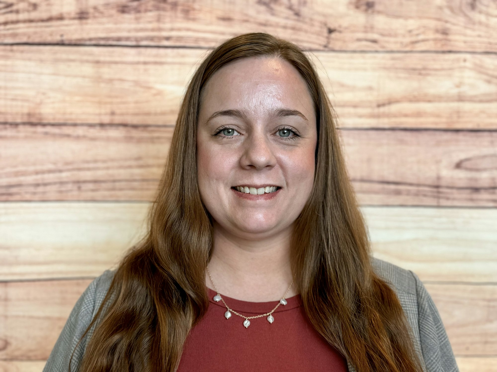 A woman smiles in front of a wooden backdrop, wearing a red top and a necklace, with long hair and a friendly demeanor.