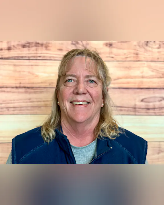 A smiling woman with long hair is wearing a navy blue vest, posing against a wood-patterned background.