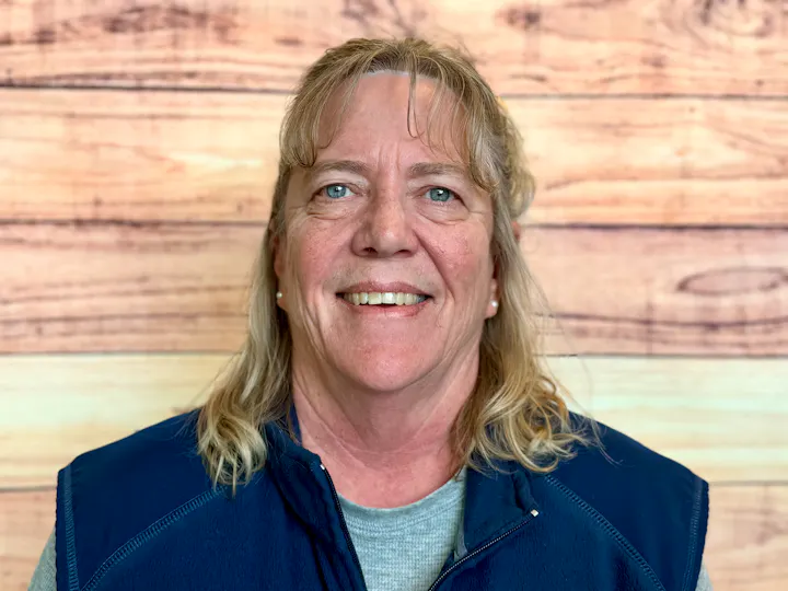 A smiling woman with long hair is wearing a navy blue vest, posing against a wood-patterned background.