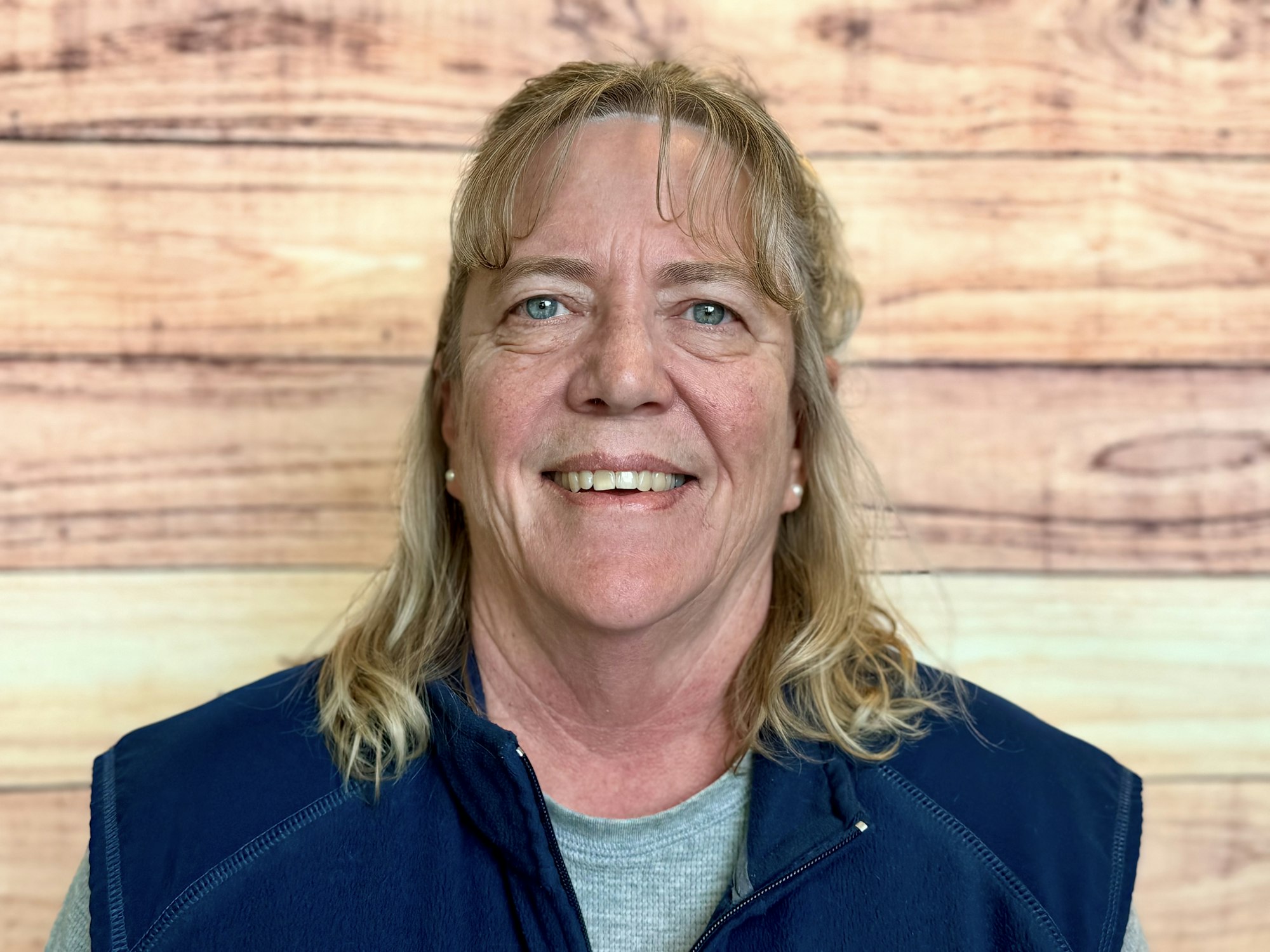 A smiling woman with long hair is wearing a navy blue vest, posing against a wood-patterned background.