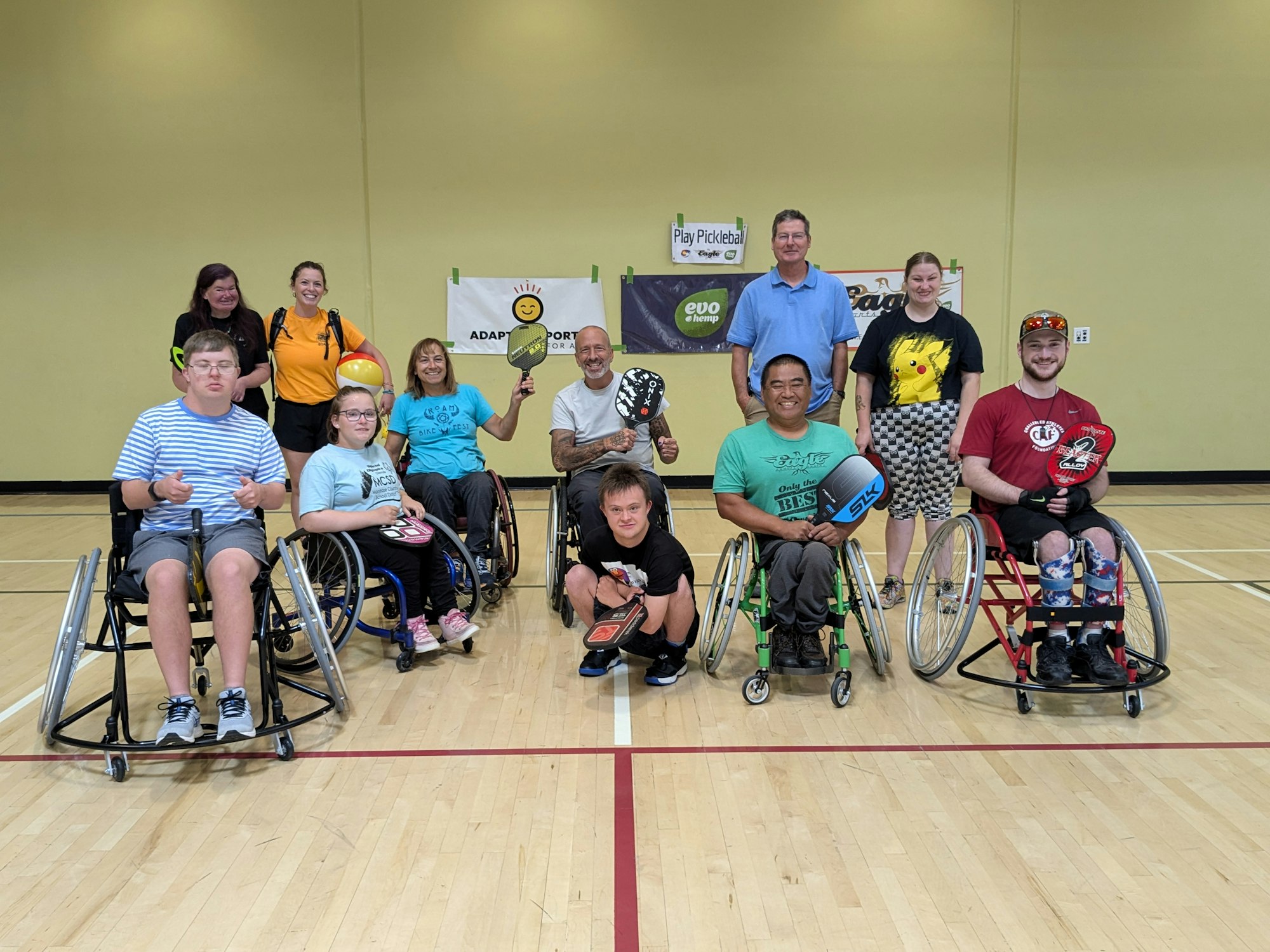 Group of smiling people with wheelchairs and pickleball paddles in a sports hall.