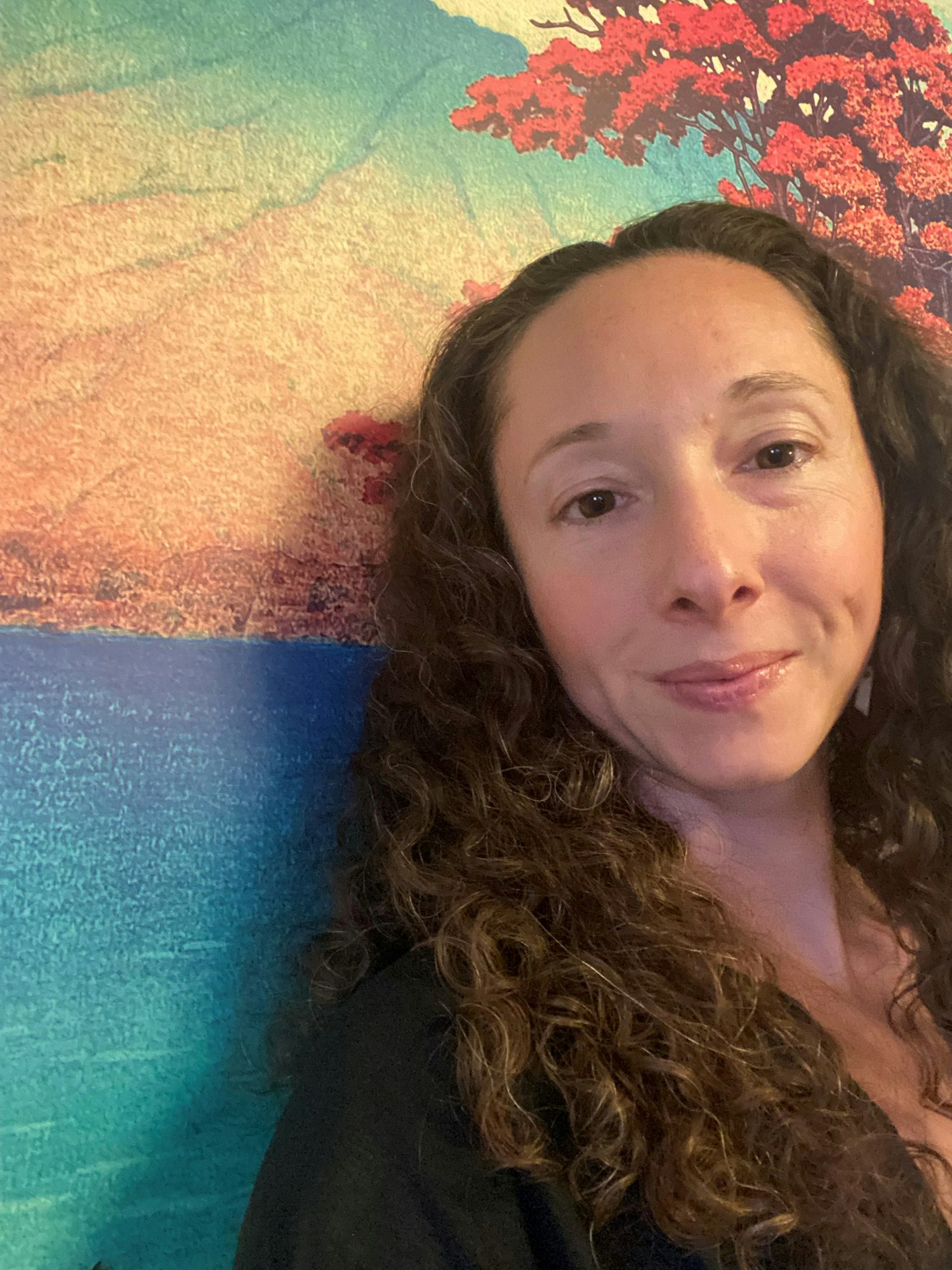 A smiling person with curly hair in front of a colorful painted canvas.