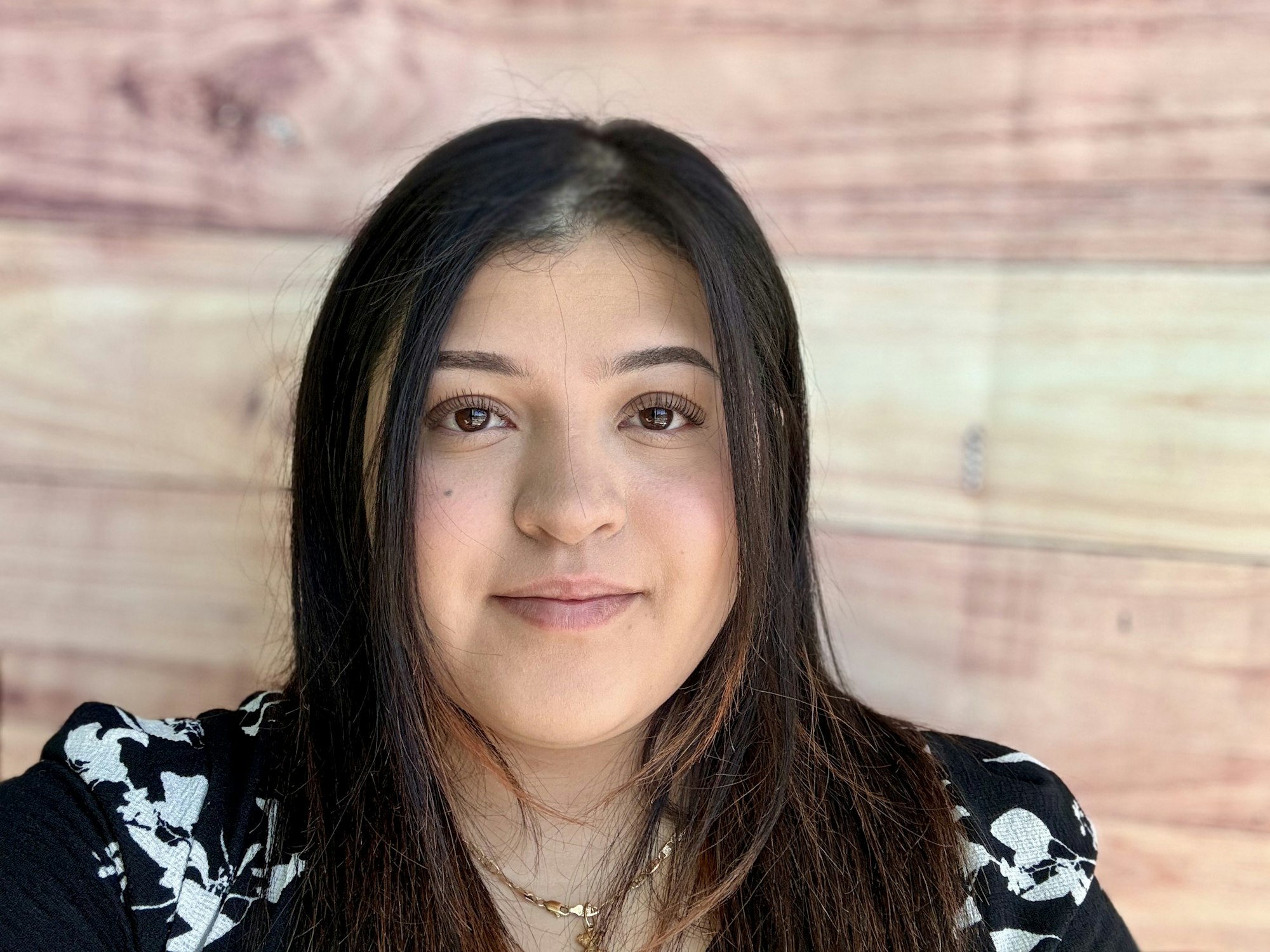 A close-up portrait of a young woman with long hair, wearing a patterned top, against a wooden background.