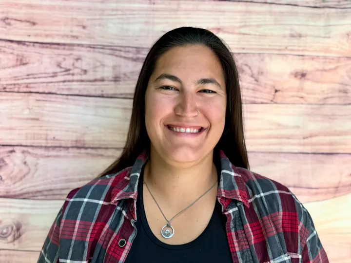 A smiling person with long hair wearing a plaid shirt stands against a wooden backdrop.