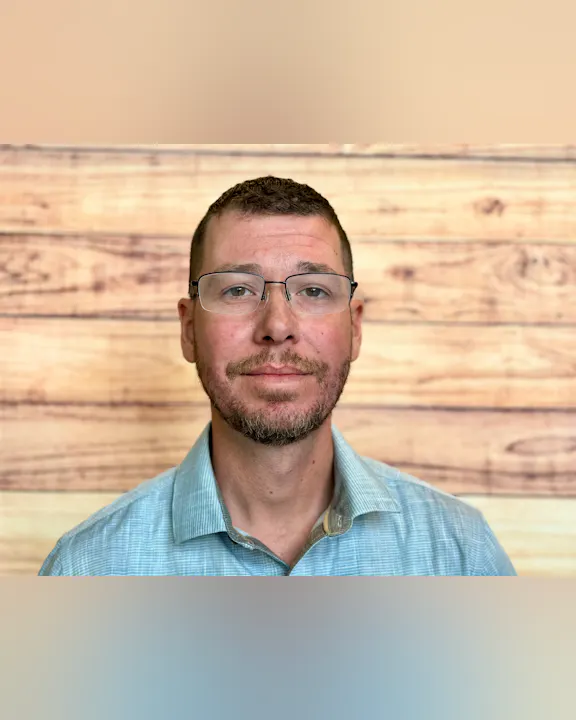 A man with glasses and facial hair is posing in front of a wooden backdrop, wearing a light blue checkered shirt.