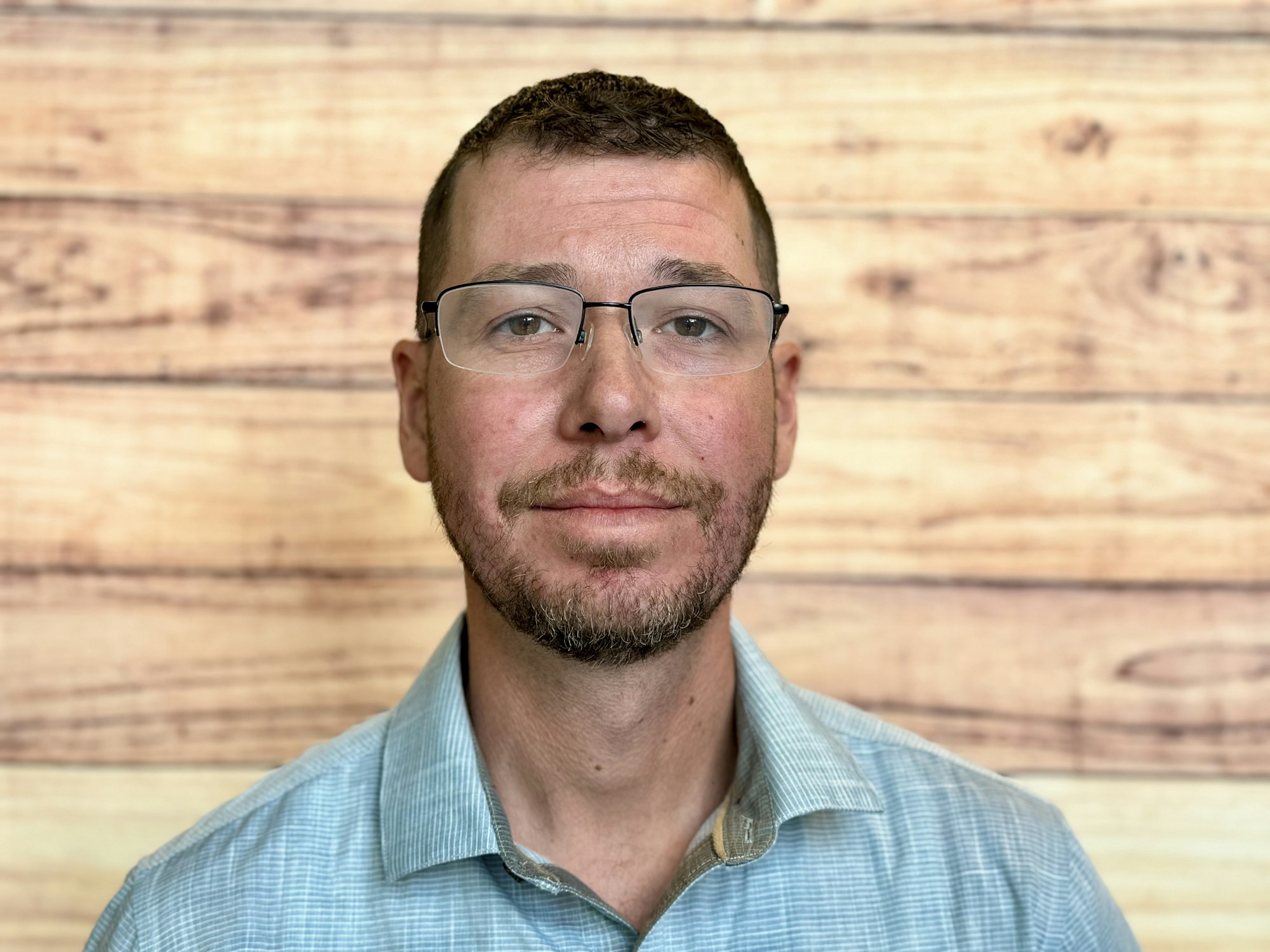 A man with glasses and facial hair is posing in front of a wooden backdrop, wearing a light blue checkered shirt.