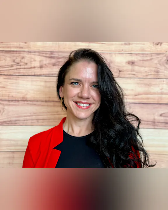 The image features a woman with long, dark hair, wearing a red blazer over a black top, smiling against a wooden backdrop.
