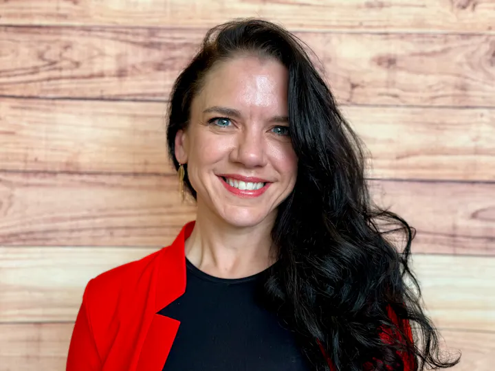 The image features a woman with long, dark hair, wearing a red blazer over a black top, smiling against a wooden backdrop.