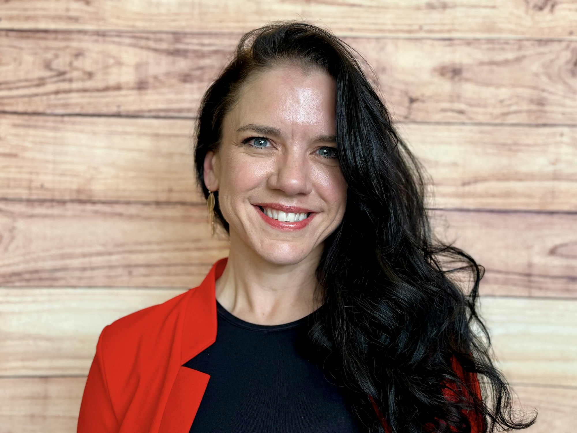 The image features a woman with long, dark hair, wearing a red blazer over a black top, smiling against a wooden backdrop.