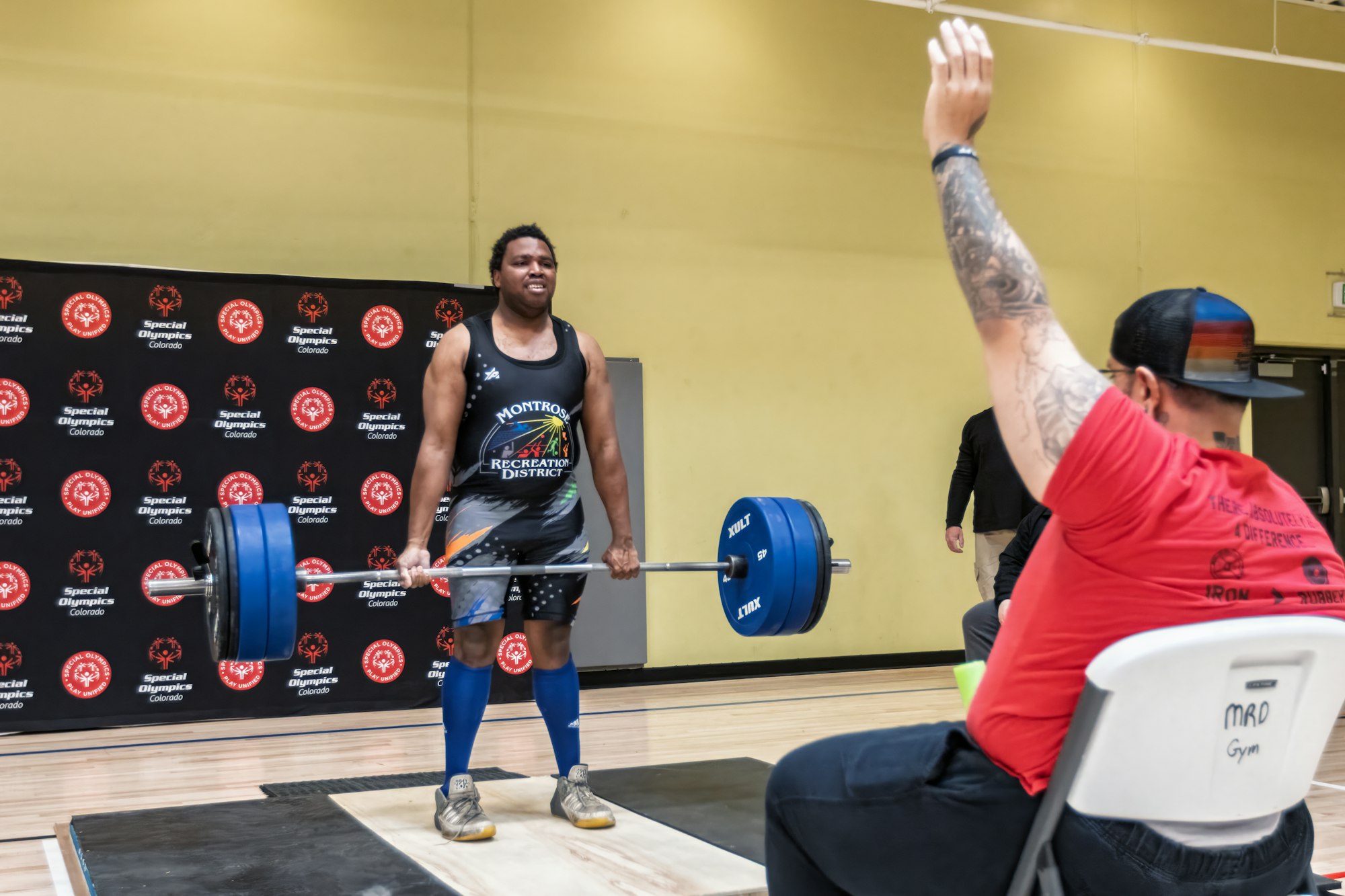 A weightlifter competes at a Special Olympics event, lifting a barbell while a judge raises their hand in approval.