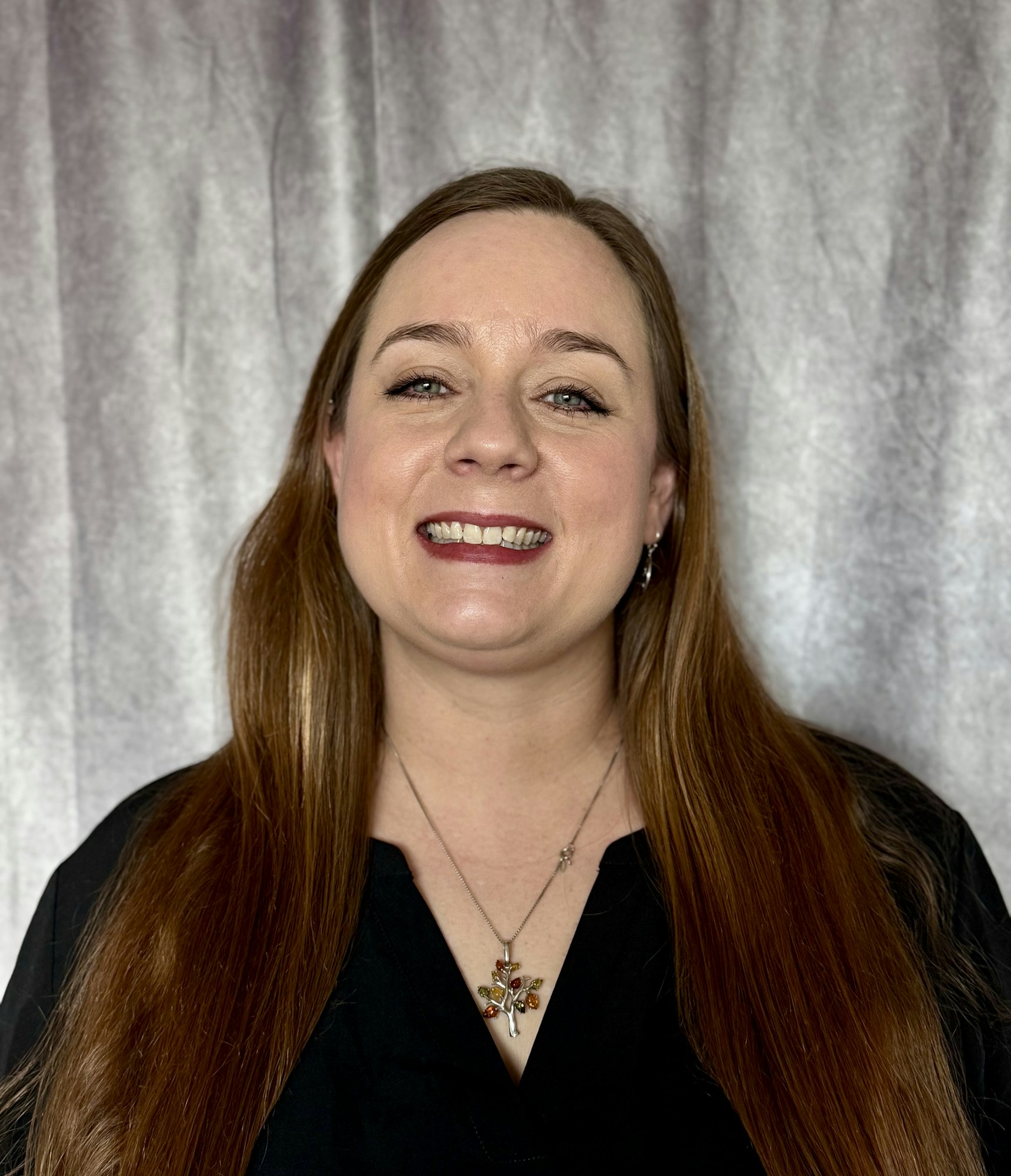 A woman with long hair is smiling while wearing a black top and a colorful necklace, set against a soft backdrop.
