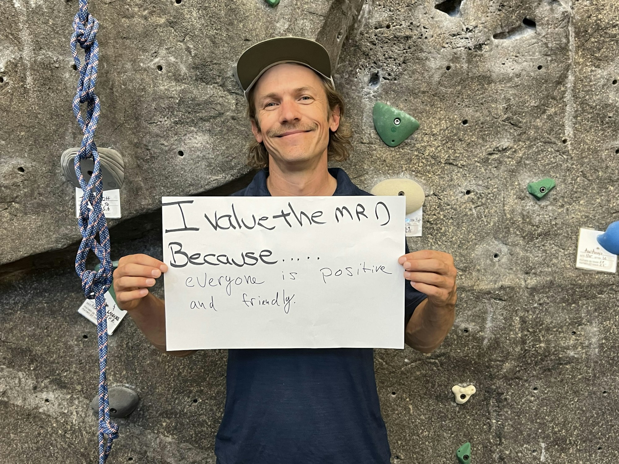 A man in a climbing gym holding a sign that says, "I value the gym because...everyone is positive and friendly."
