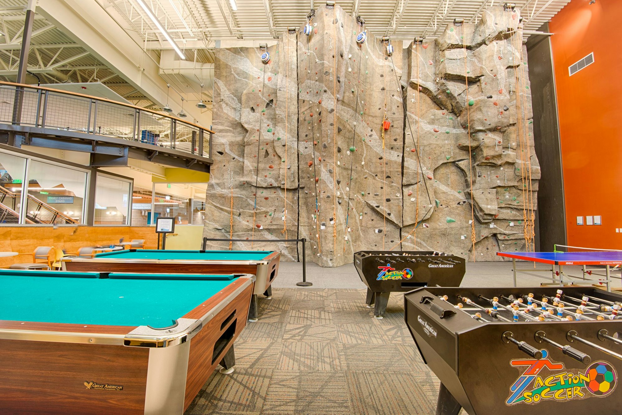 The image features a recreational area with a climbing wall, pool tables, foosball tables, and a vibrant interior design.