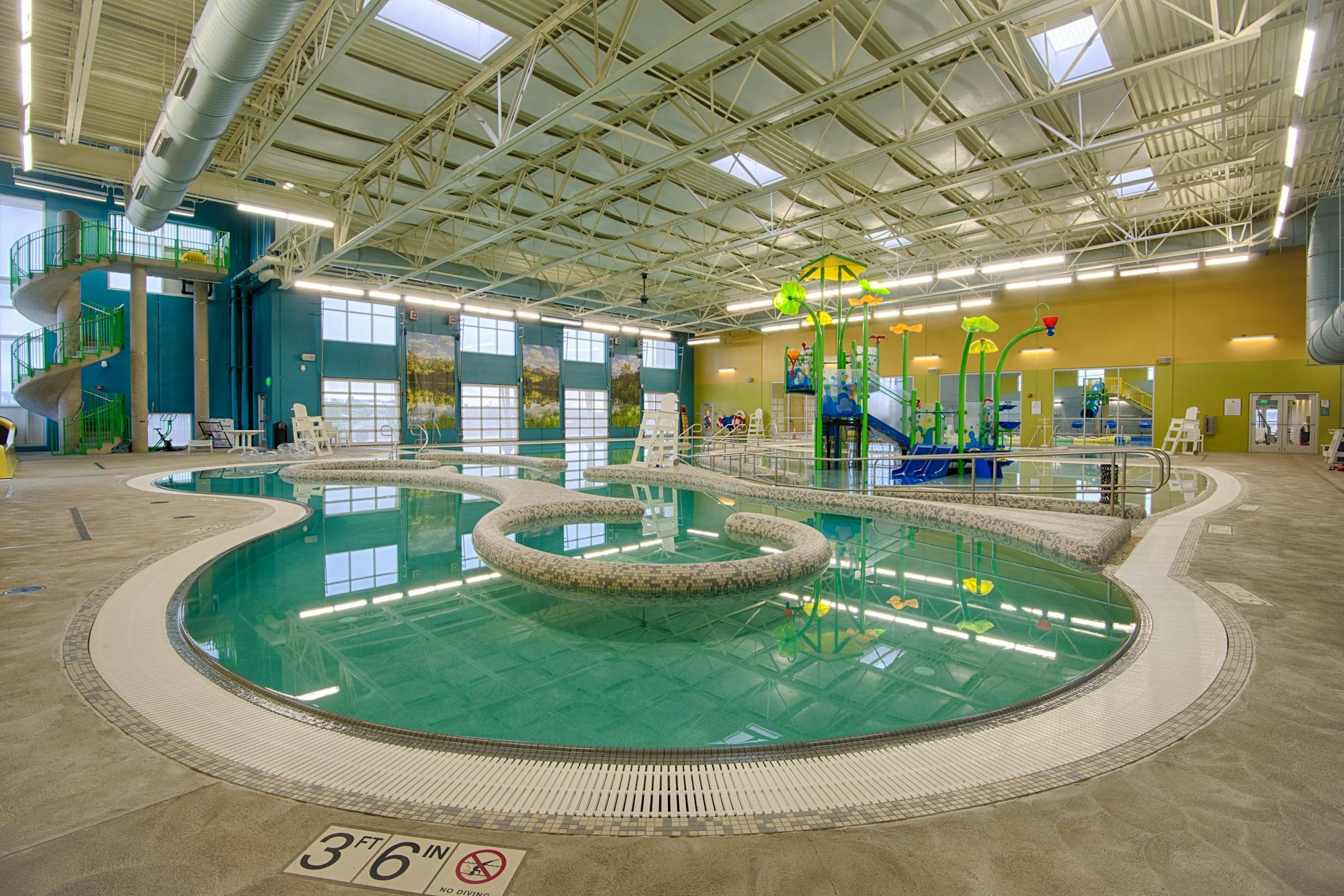An indoor swimming pool area featuring a slide, play structures, and a winding layout with lounge chairs and safety signs.