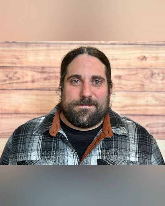 The image features a man with a beard, wearing a plaid shirt, set against a wooden background.