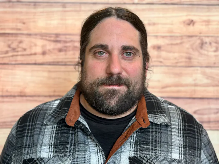 The image features a man with a beard, wearing a plaid shirt, set against a wooden background.
