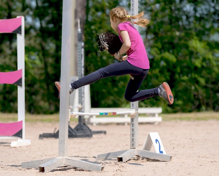 A girl in a pink shirt is jumping over an obstacle, holding a hobby horse, in an outdoor setting.