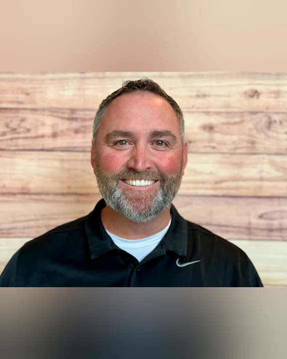 The image shows a smiling man with a beard, wearing a black shirt, set against a wooden background.