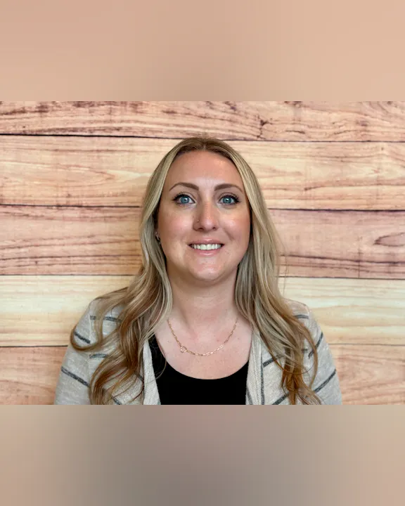 A woman with long, blonde hair is smiling against a wooden background. She's wearing a striped cardigan and a black top.