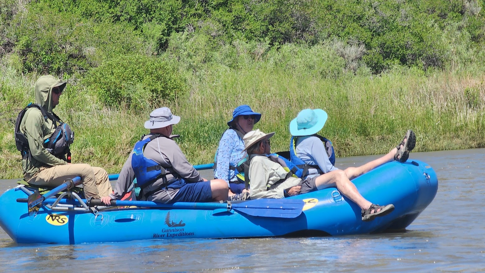 Five people rafting on a river in an inflatable boat, surrounded by greenery.