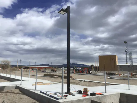 An outdoor construction site with a newly installed light pole, concrete foundations, and machinery in the background under a cloudy sky.