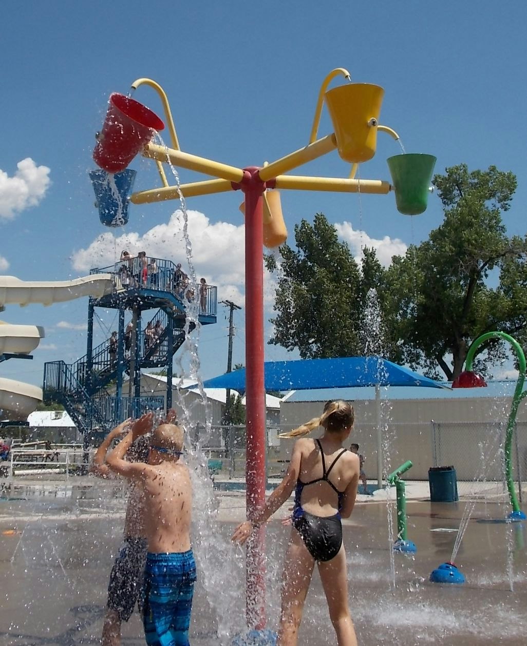 Children playing in a splash park under a water feature with tipping buckets.