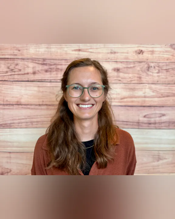 A smiling person with glasses stands in front of a wooden backdrop, wearing a brown shirt over a black top.