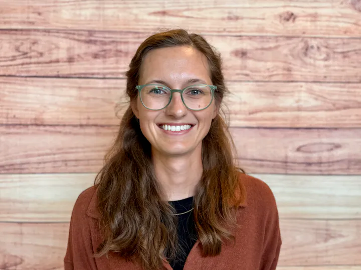 A smiling person with glasses stands in front of a wooden backdrop, wearing a brown shirt over a black top.