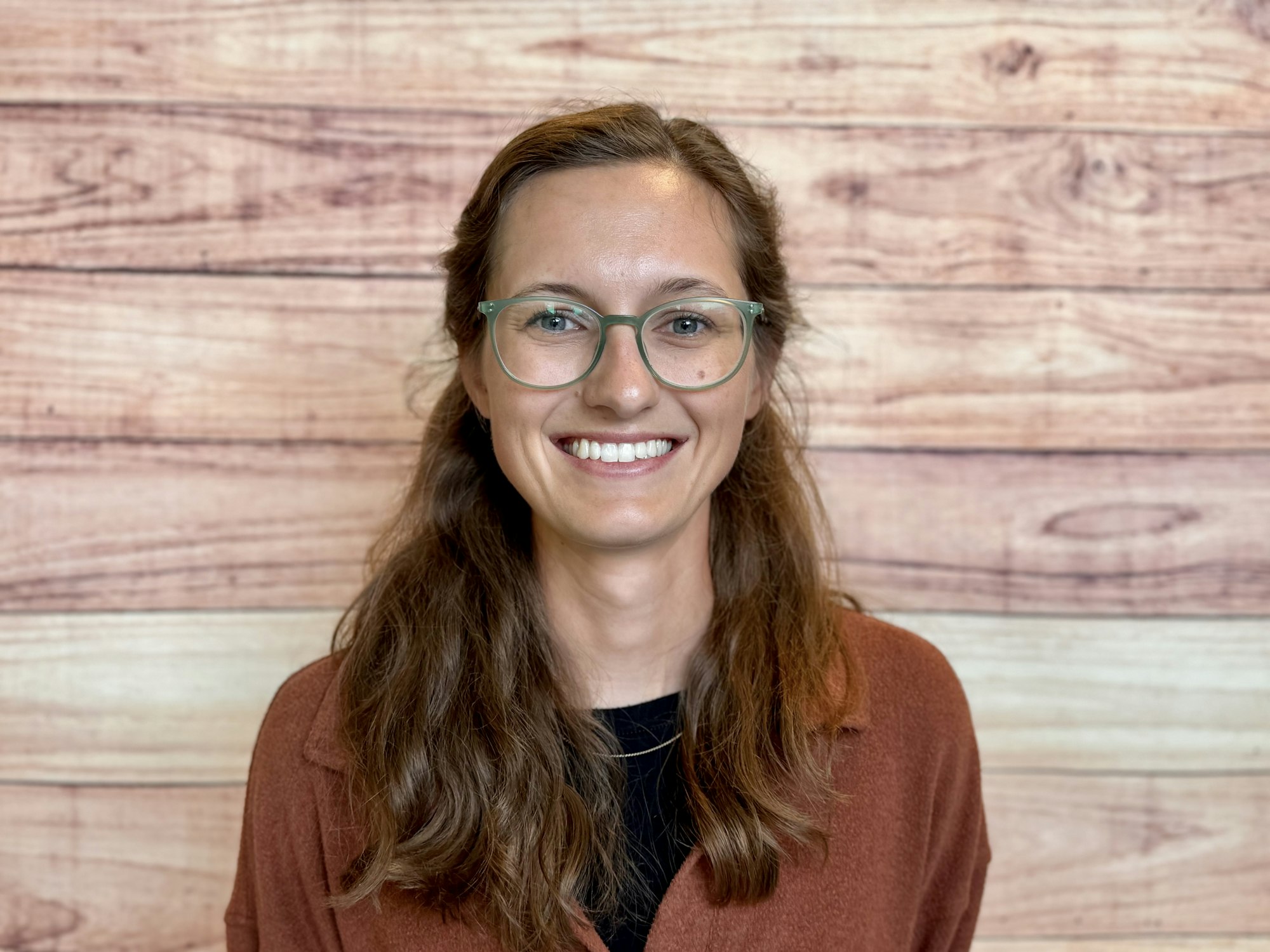 A smiling person with glasses stands in front of a wooden backdrop, wearing a brown shirt over a black top.