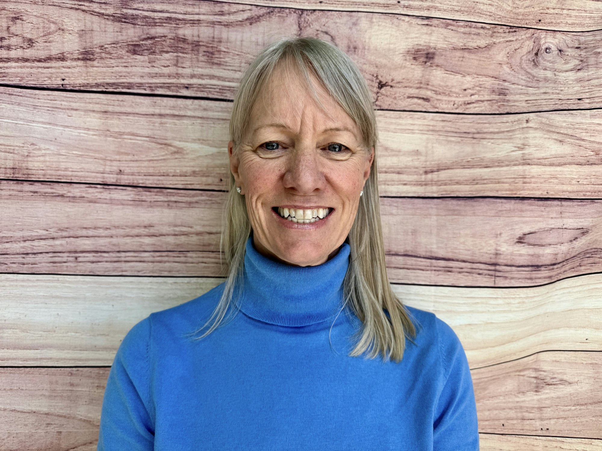 A smiling person with long, light hair, wearing a blue turtleneck, stands against a wooden textured background.