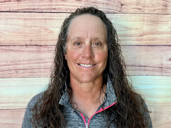A person with long, curly hair smiles in front of a textured wooden backdrop. They wear a gray zip-up jacket.