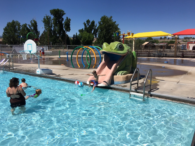 A colorful children's pool with a frog slide, people swimming, and play structures.