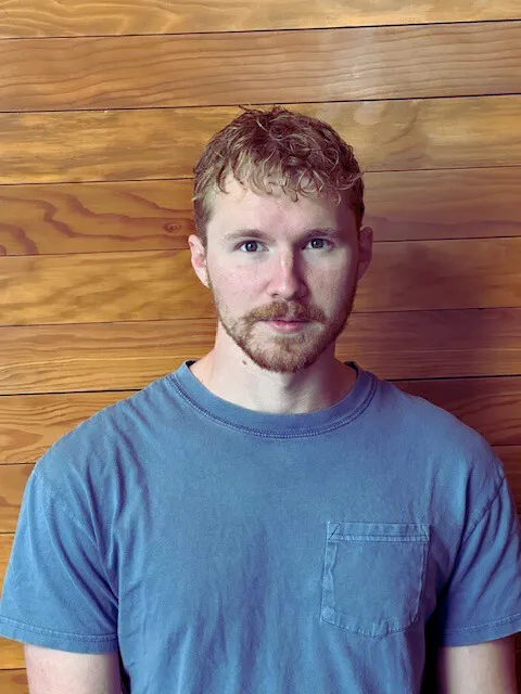 A person with short, curly hair and a beard stands against a wooden wall, wearing a blue shirt.