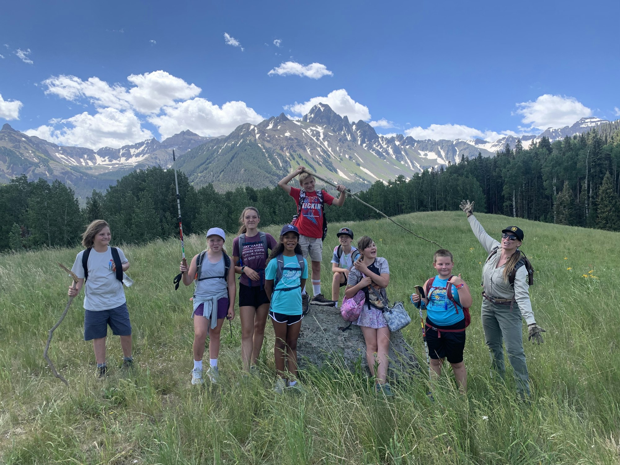 A group of people, including children and a leader, pose with sticks in a grassy field with mountains in the background.