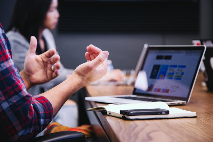 A person is engaged in discussion at a table with a laptop, notebook, and phone, while another person works in the background.