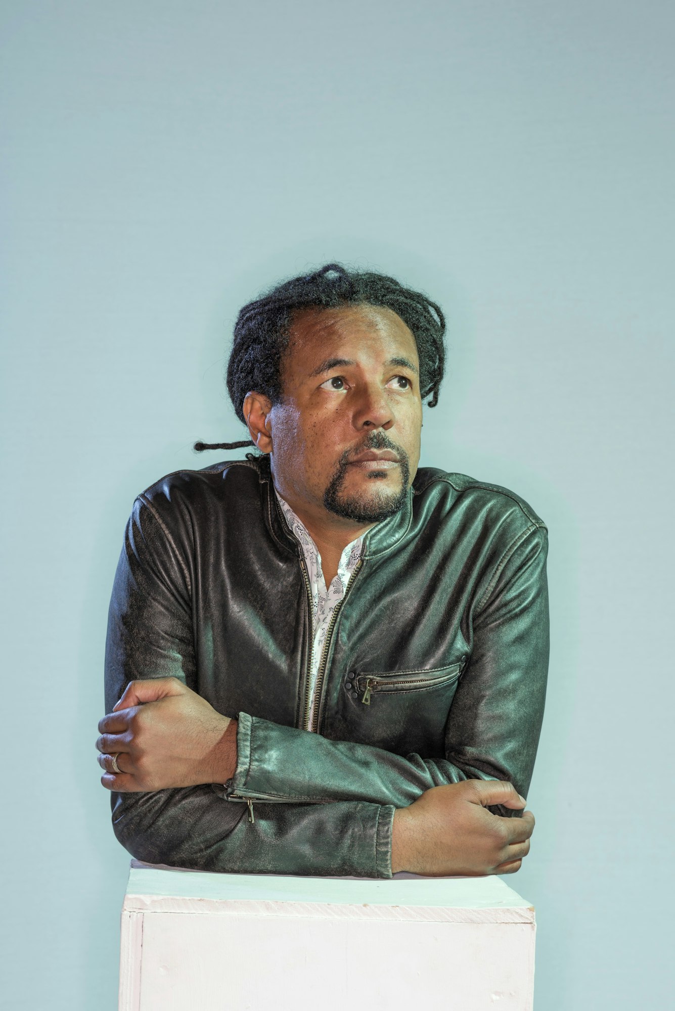 A man with dreadlocks is wearing a leather jacket, looking thoughtfully upwards against a light background.