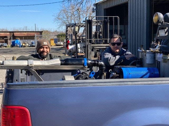 Two people stand behind a truck with equipment, near a warehouse under sunny skies.