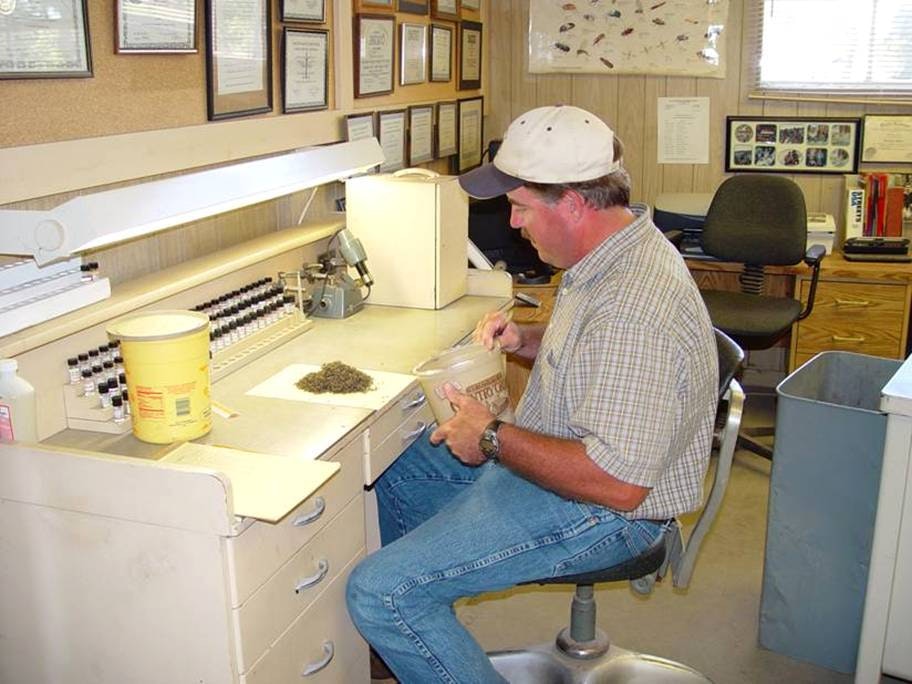 A person analyzing a sample at a desk, surrounded by lab equipment and framed certificates on the wall.