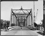 A black and white image of a metal truss bridge over a road, with construction equipment visible in the background.