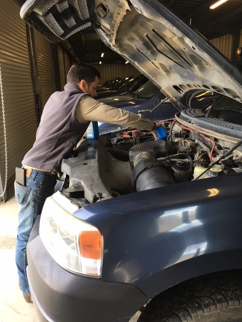 A person is working under the open hood of a vehicle in a garage.