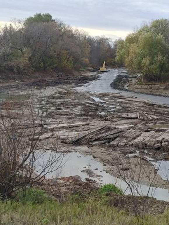 A construction vehicle on a muddy, partially dried riverbed surrounded by trees.