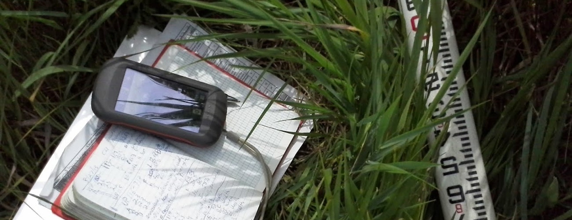 A GPS device on a notebook with handwritten notes, a measuring tape, and surrounding grass.