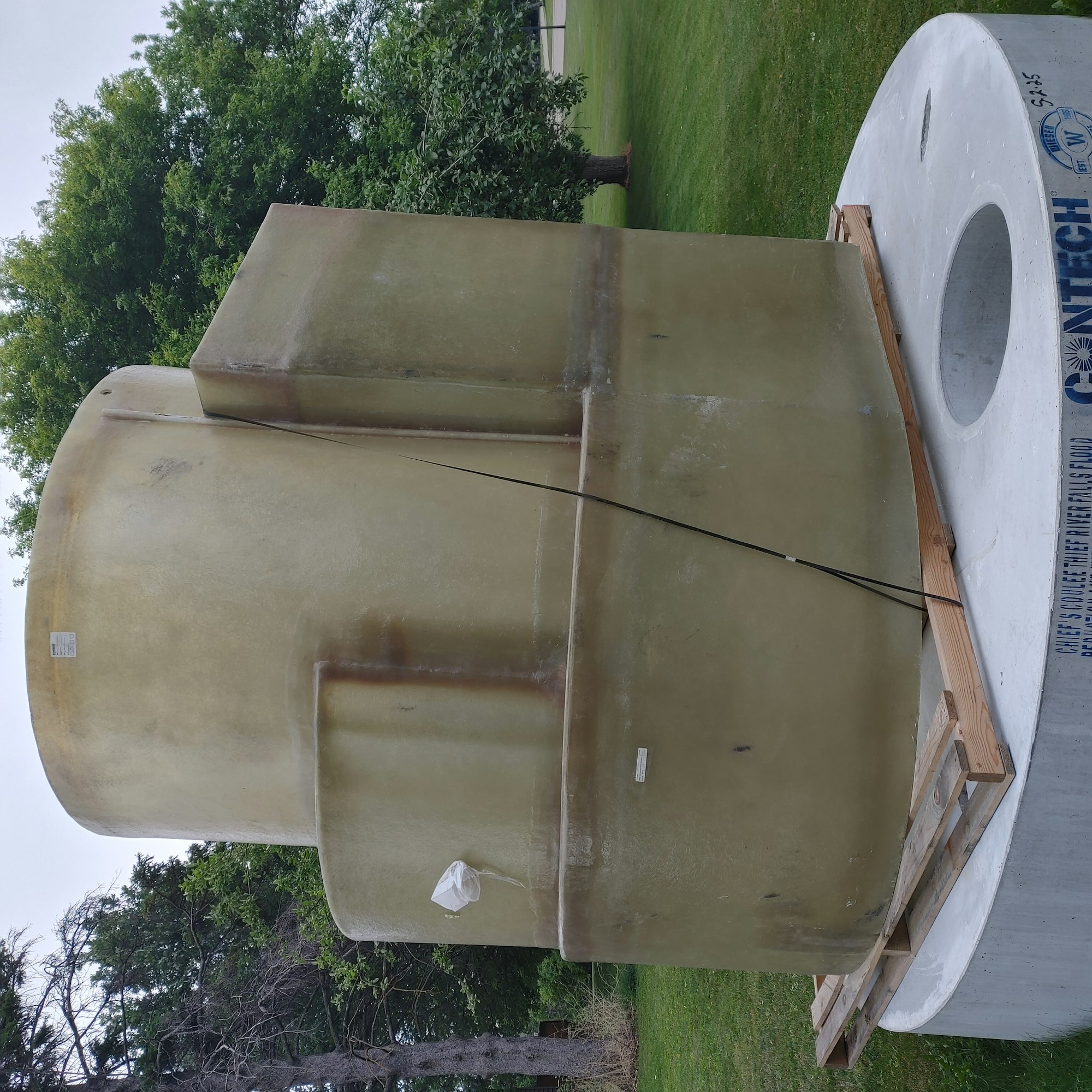 A large cylindrical structure is mounted on a concrete base outdoors, with trees and grass in the background.