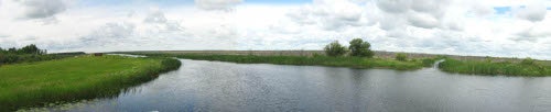 A serene landscape with a river, grassy banks, and a cloudy sky.